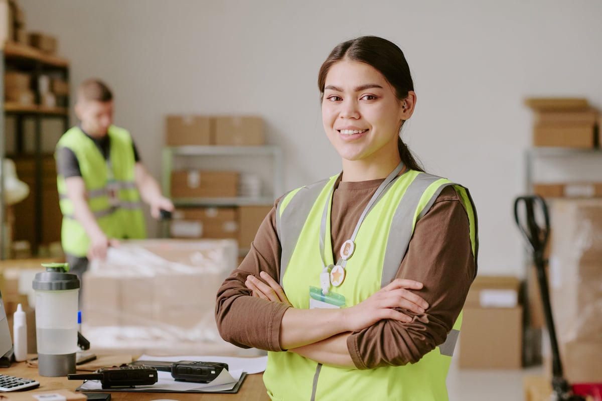 Smiling employee working in a warehouse while unpacking boxes and wearing a hi-vis vest in a storage area, visible carton boxes and working equipment in background