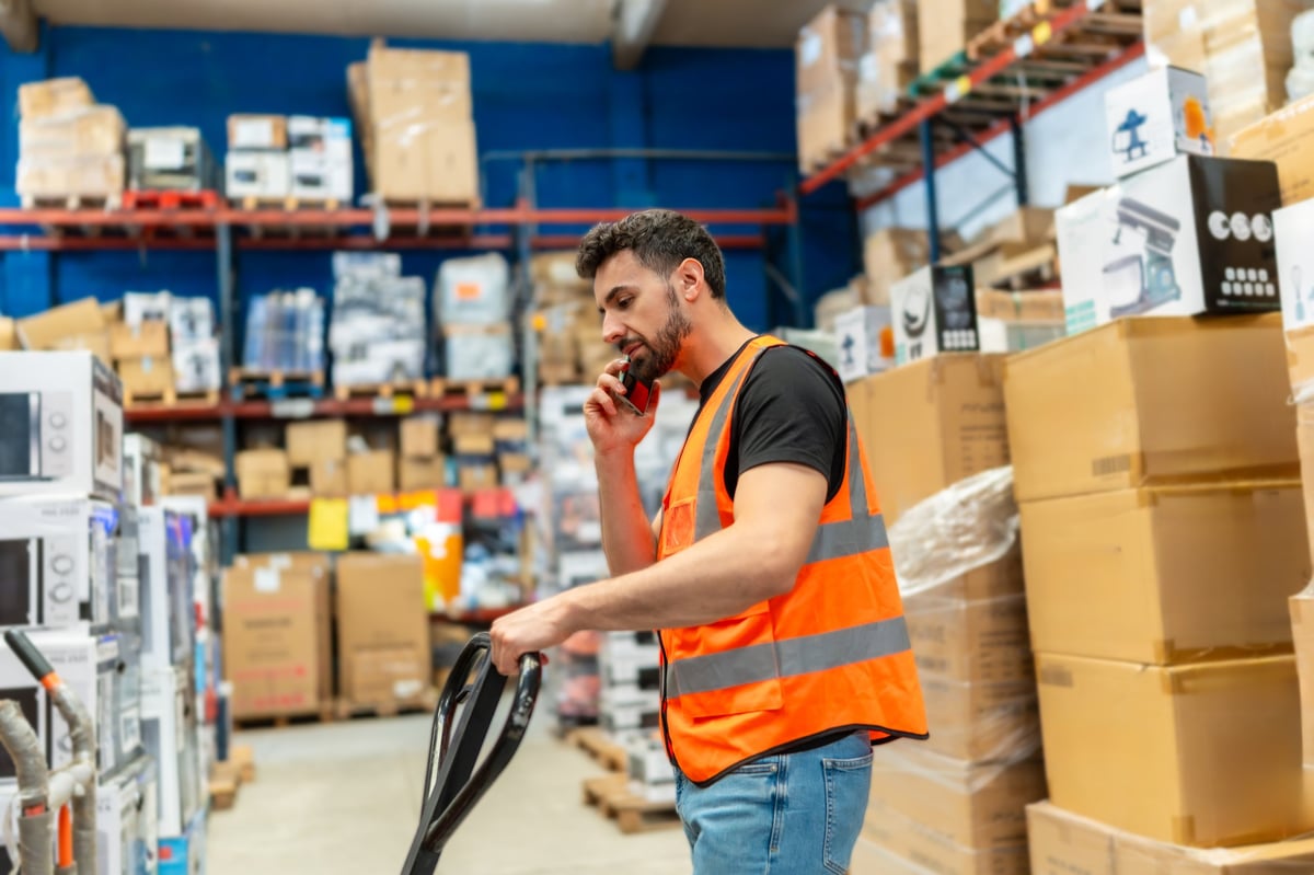 Warehouse staff member wearing high visibility vest talking on a mobile phone while pulling a pallet jack down an aisle in a busy distribution center or large retail store